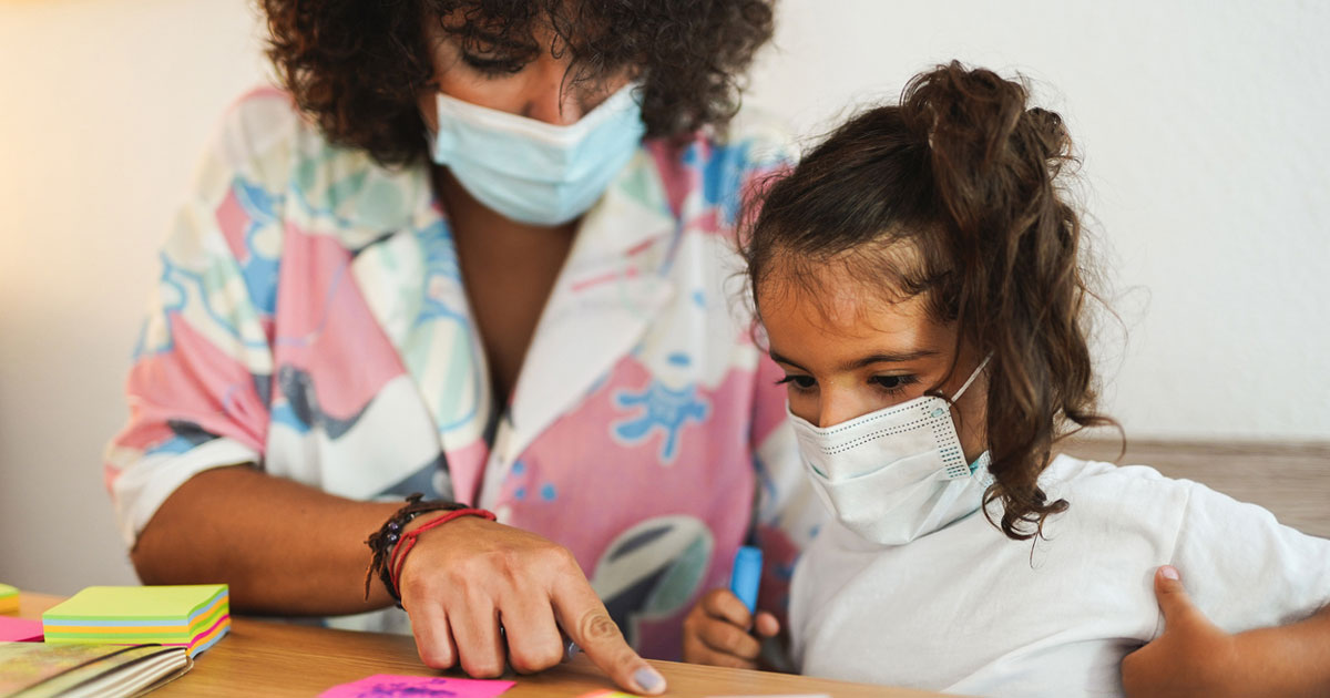 An adult and child, both wearing surgical masks, at a desk with colorful paper.
