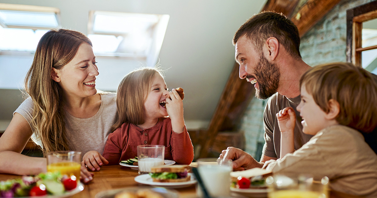 A family of four sits around a wooden table in a sunlit home, smiling and eating breakfast together.