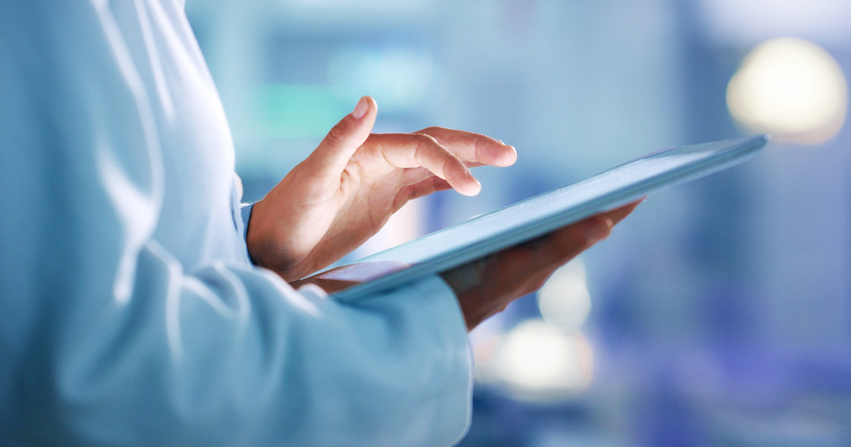 A person in a white coat uses a tablet device, pointing with a finger. The background is softly blurred, suggesting a professional, clinical setting.
