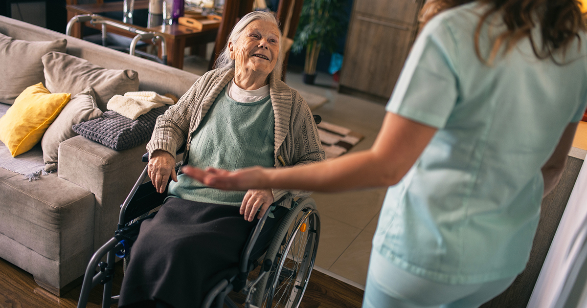 An older woman seated in a wheelchair smiles while speaking with a healthcare worker in scrubs during a home visit.