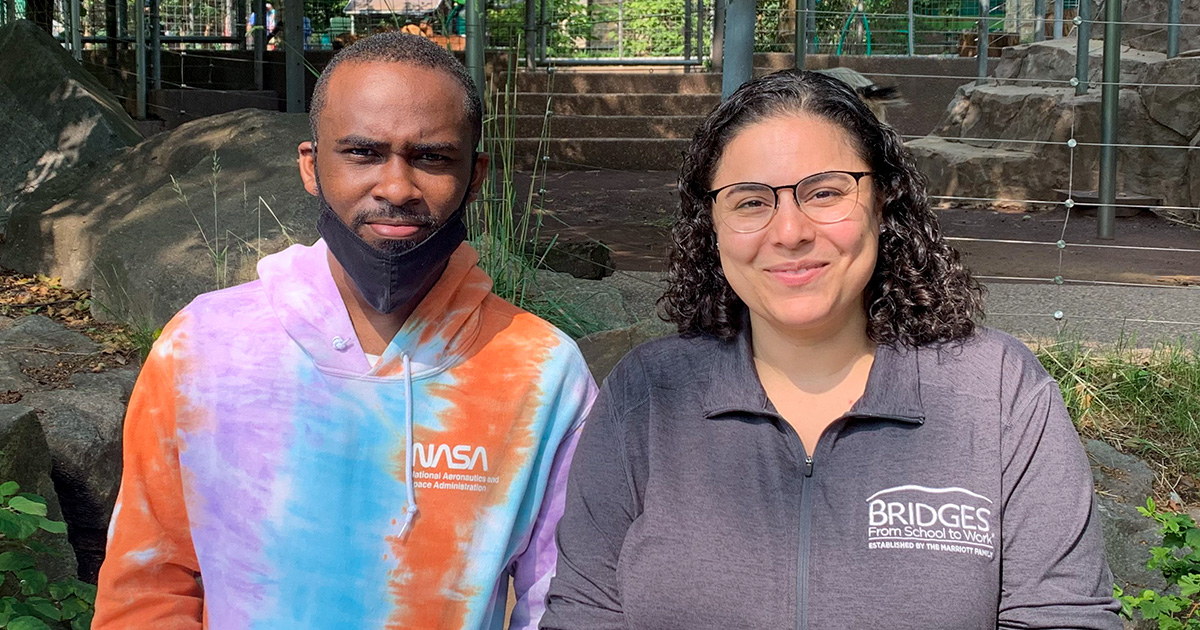 Two people smile at the camera in front of a park, one wearing a shirt with the Bridges logo