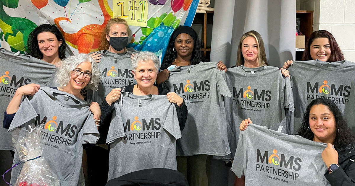 A group of people stand smiling at the camera, each holding up a t-shirt with the MOMS logo on it