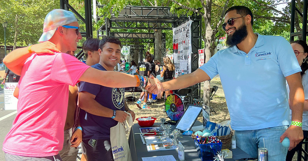 Two people shake hands over a table filled with pamphlets in a busy public outdoor area