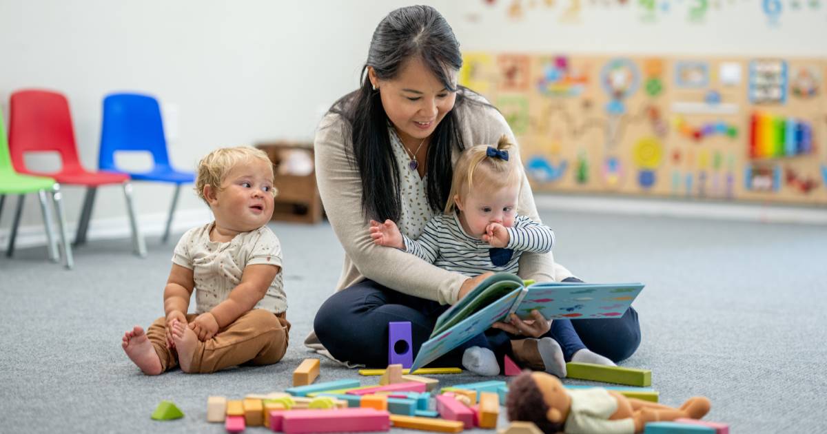 Two infants in a classroom looking at a book with their teacher or caregiver