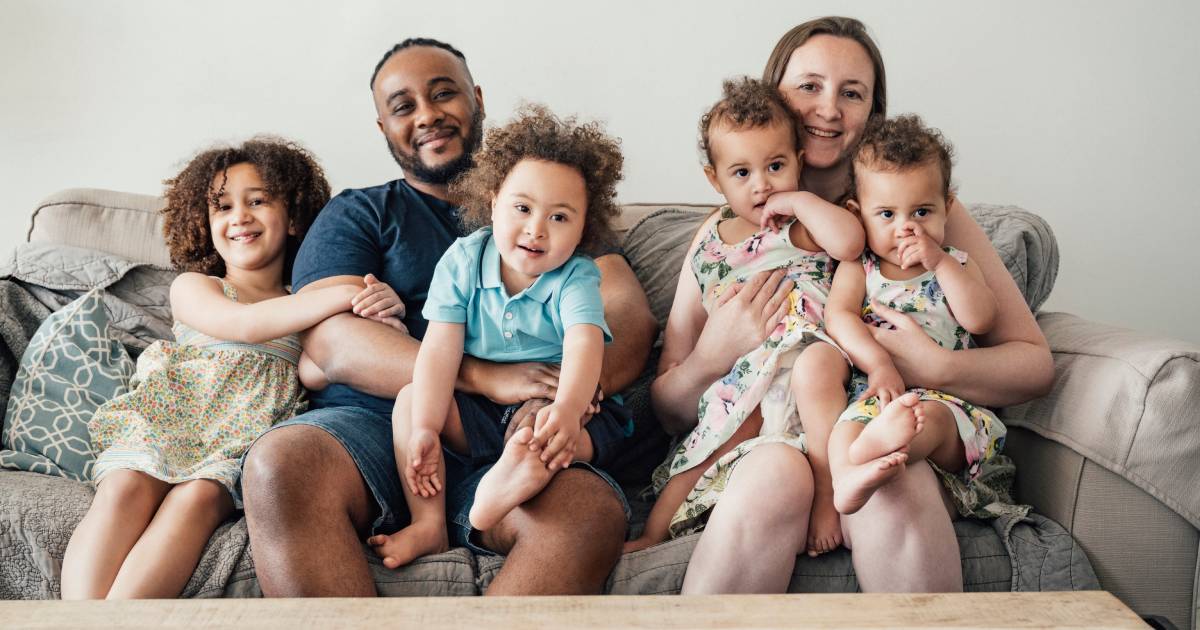 Two parents with four young children sitting on their couch together and smiling