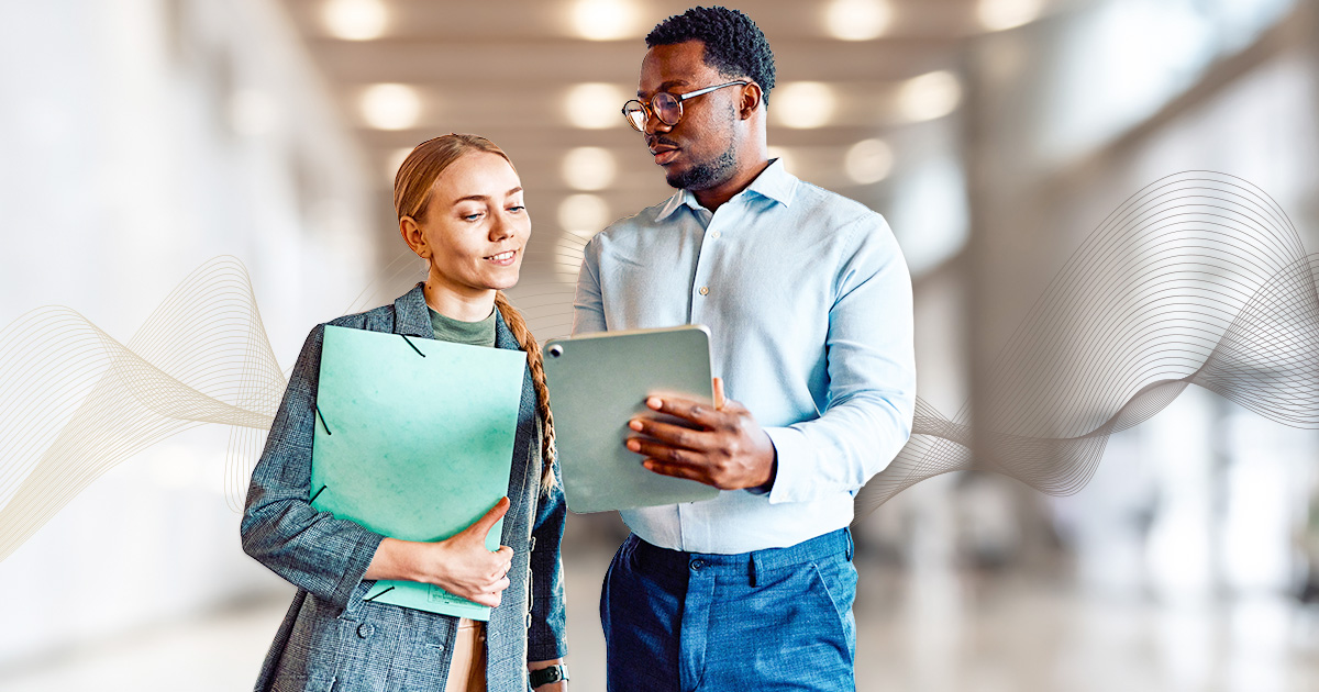 A man and a woman in professional attire discuss something on a tablet in a modern, bright hallway. She holds a teal folder, indicating collaboration.