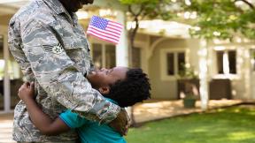 Side view close up of a young adult African American male soldier in the garden outside his home, embracing his young son, who is looking up at him smiling and holding a US flag