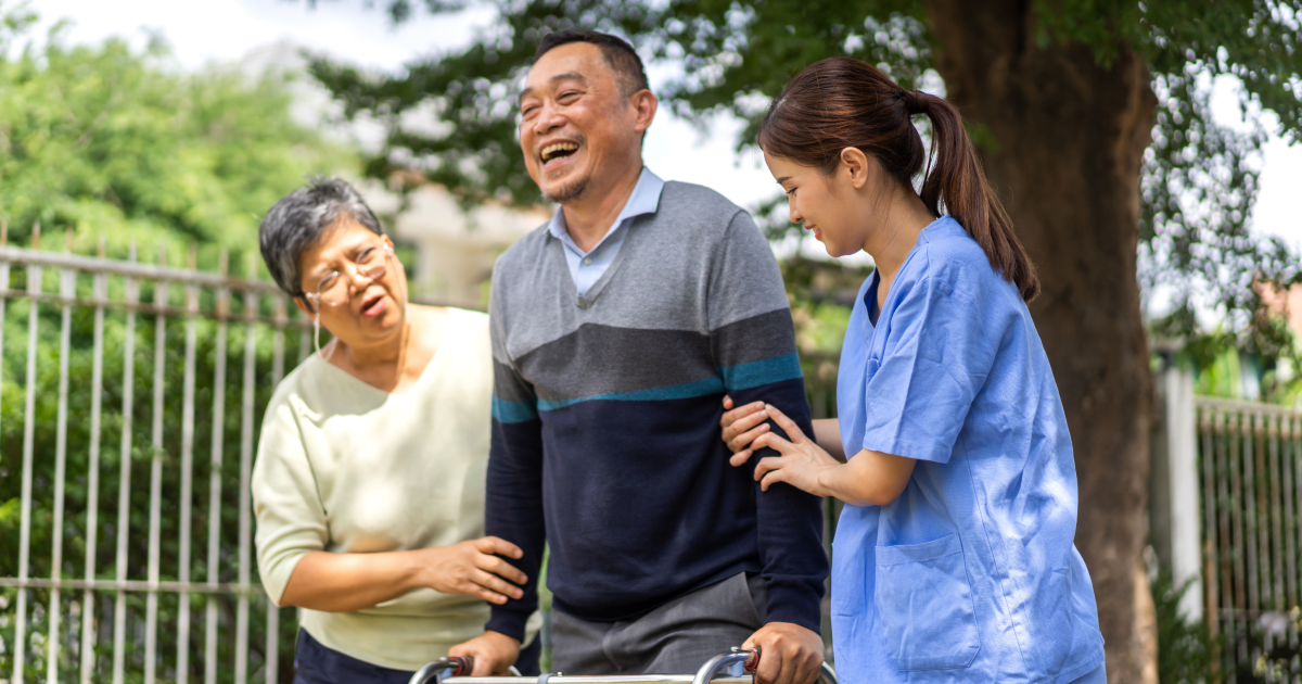 A smiling man using a walker with assistance of caregivers.