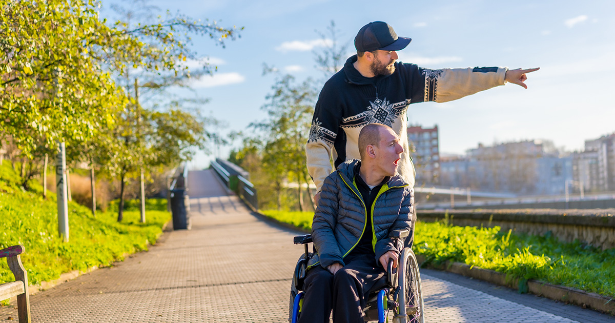 Man pushing young man in a wheel chair on a city sidewalk by a park