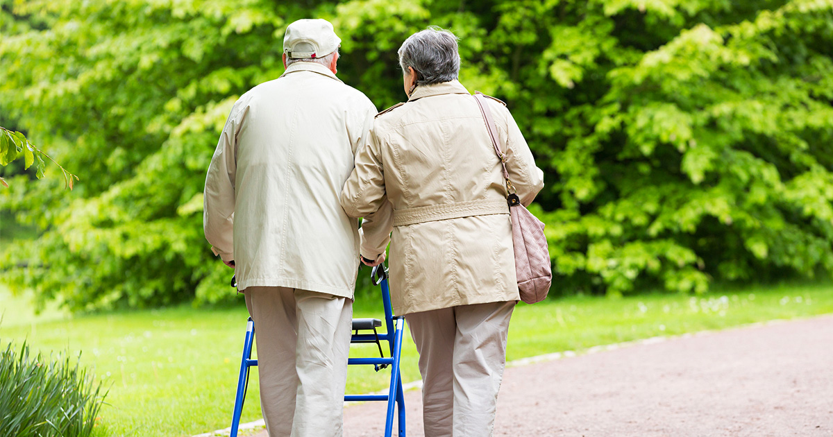 Older couple walk together along a park path, with one person using a walker for support.
