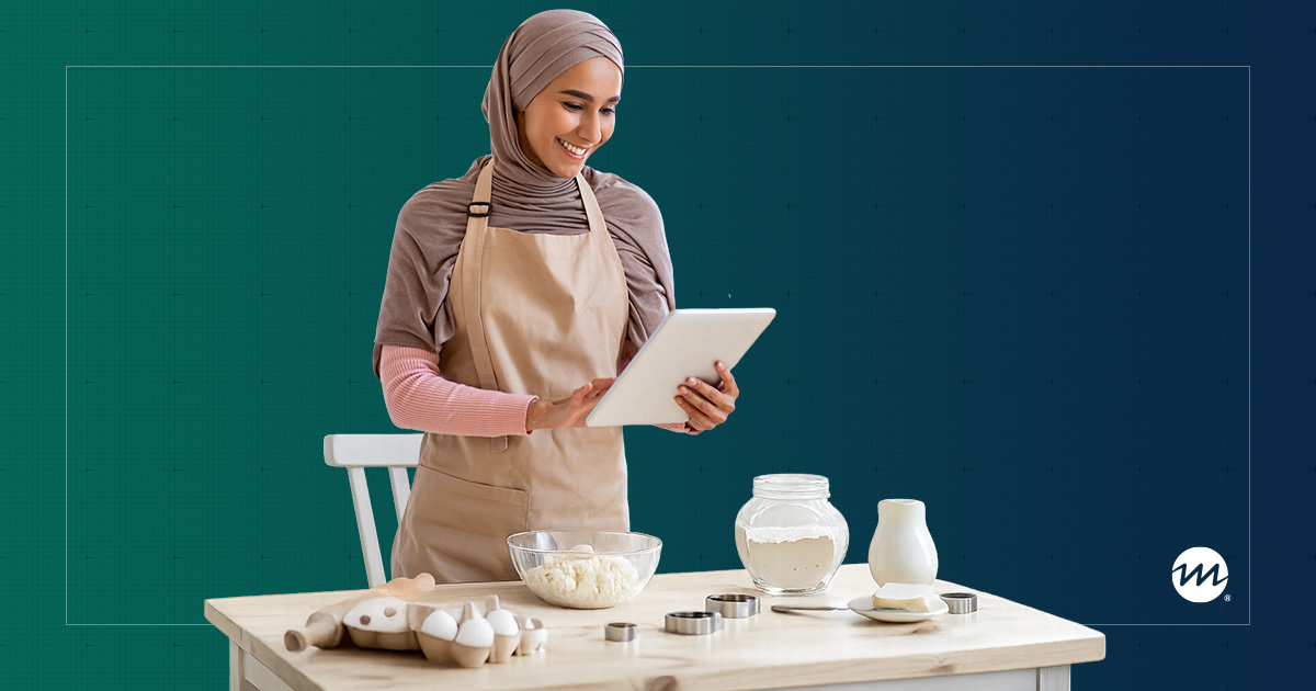 A woman in a hijab and apron smiles while using a tablet, standing at a kitchen table with baking ingredients
