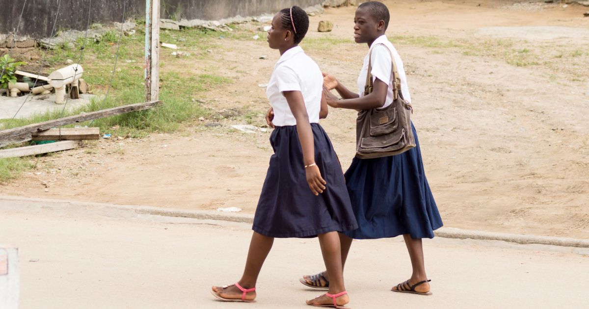 Two students in school uniforms, one wearing a white shirt and blue skirt, the other a white shirt and navy blue skirt, walking together on a paved path.