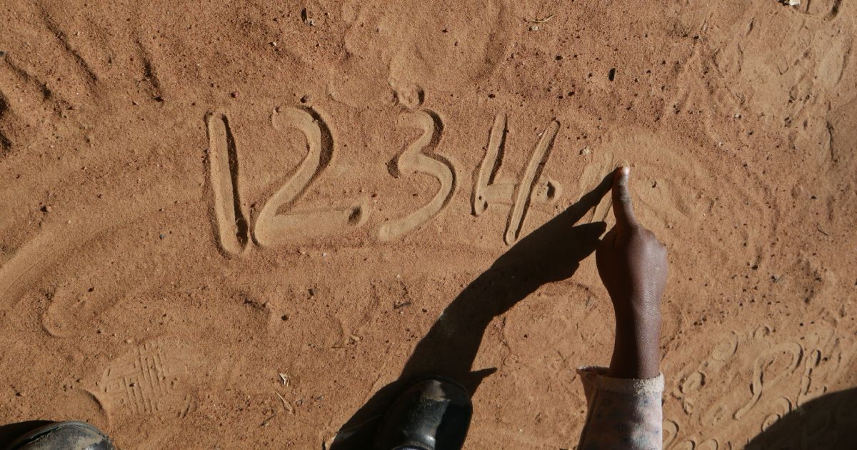 A child's hand points to numbers "1234" drawn in the sand