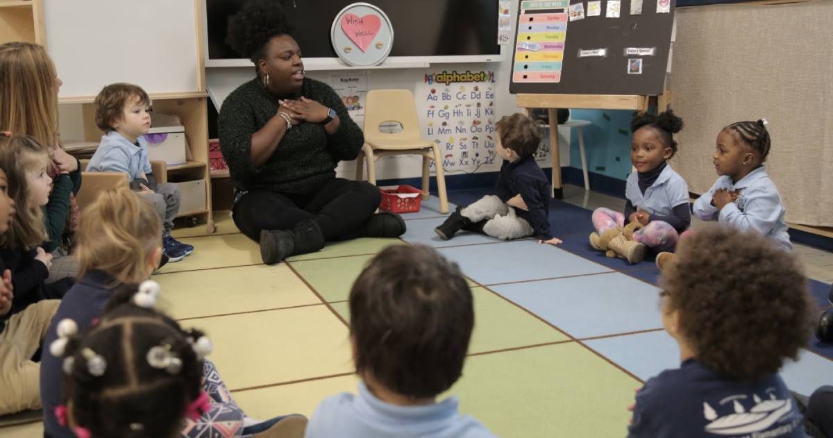 A teacher sits on the floor with a group of young children in a classroom, engaging in an activity. Behind them, an alphabet chart and posters are displayed on the wall.