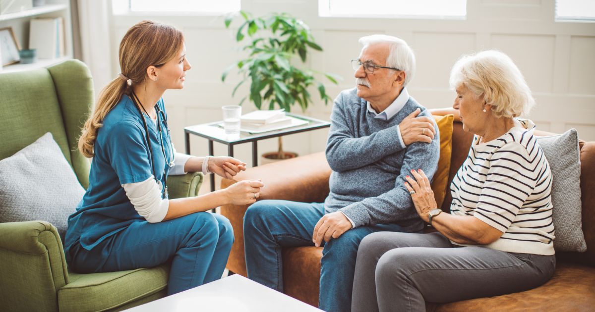 A nurse talking to an elderly couple, with the man holding his shoulder as they sit on a couch in a bright living room.