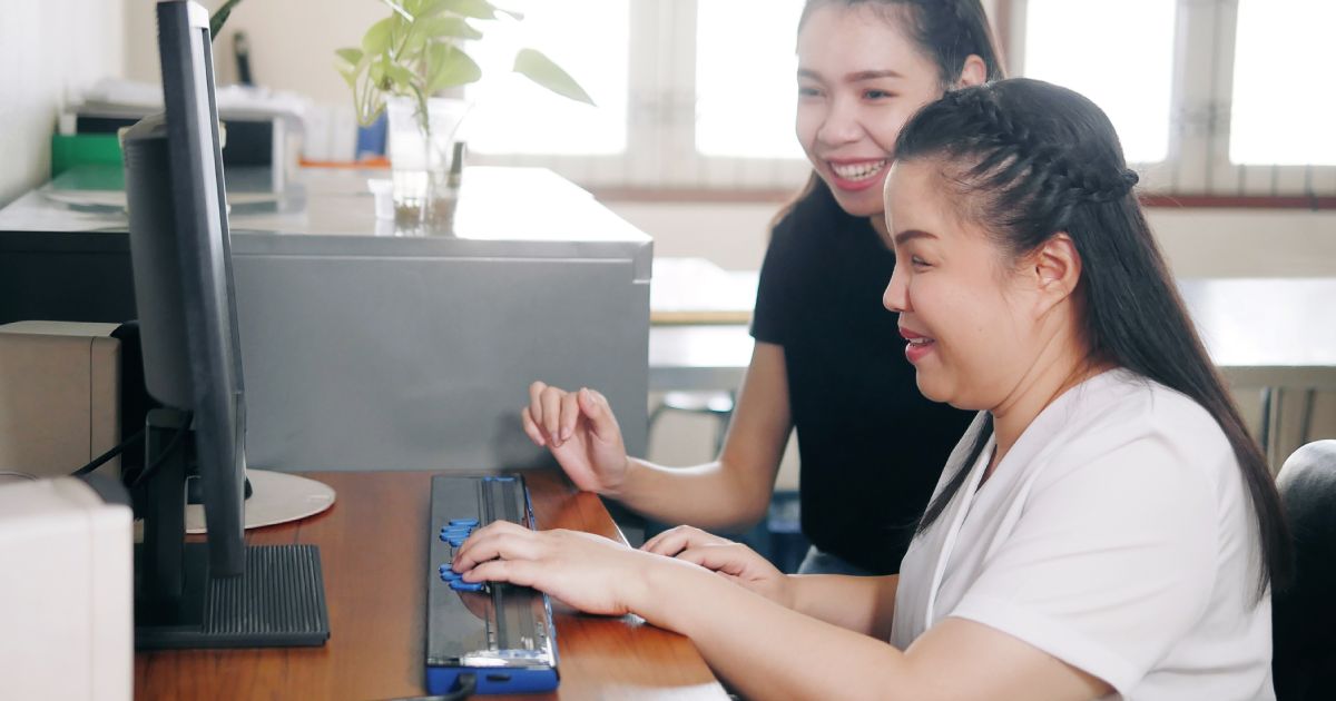 Happy Asian women co-workers in office workplace including person with blindness disability using computer with refreshable braille display assistive device.