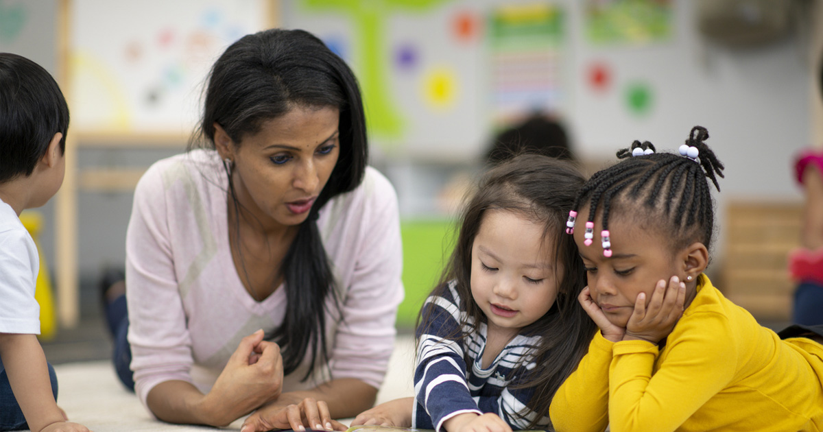 An ethnic teacher is laying on the floor with a group of students. The educator is reading to the preschool kids. They are in a clean and modern classroom. One student is pointing to a page of the book and asking a question.