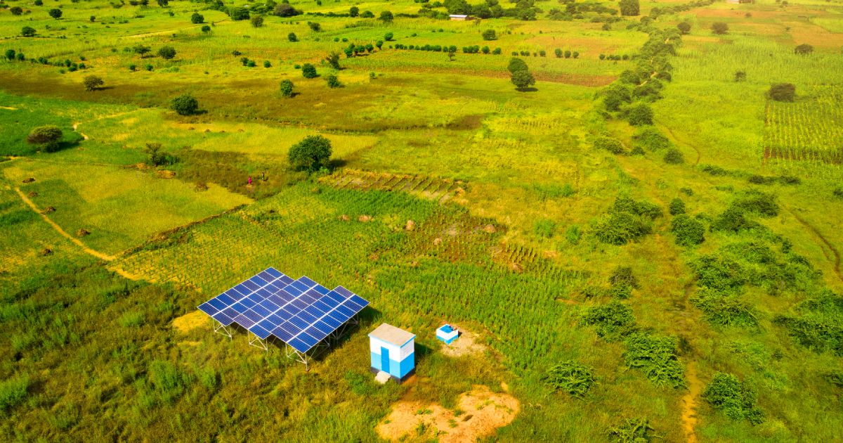 Aerial view of a solar panel installation and small building in a green, rural landscape with fields and sparse trees.