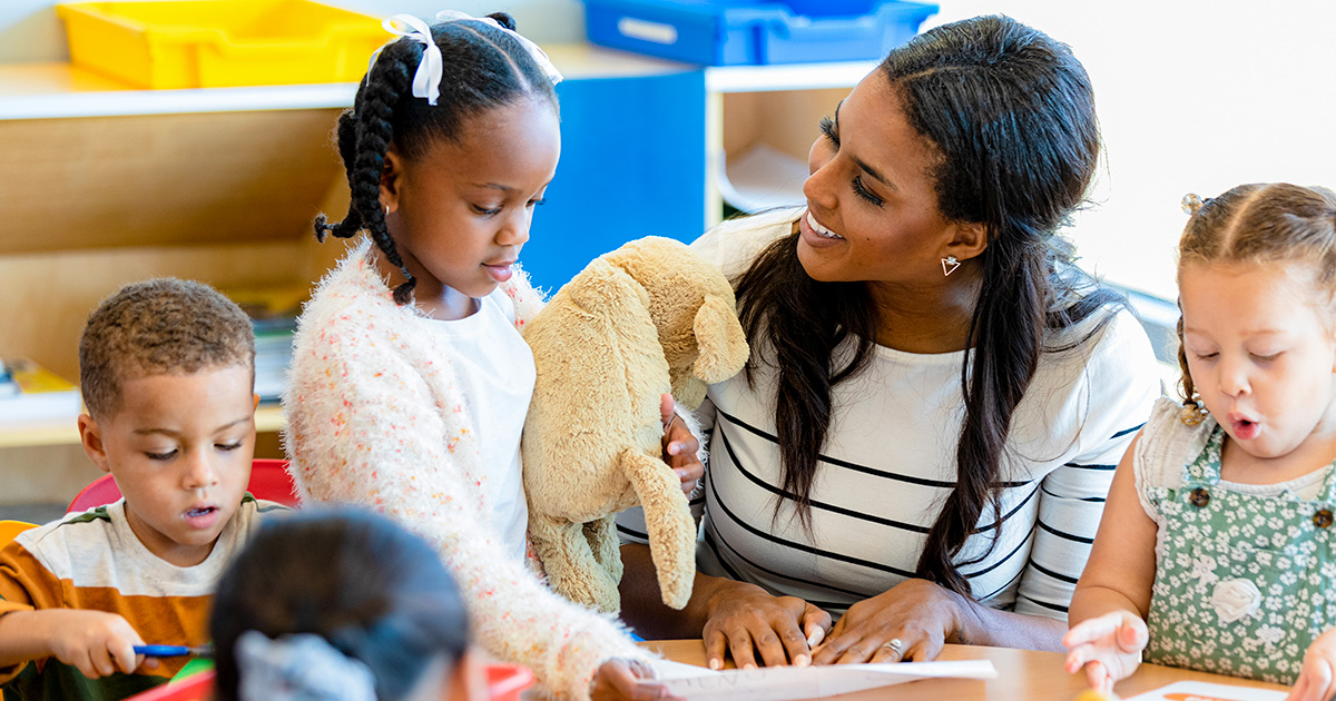 Teacher engages with young children during a hands-on classroom activity, holding a stuffed animal while students work at a table.