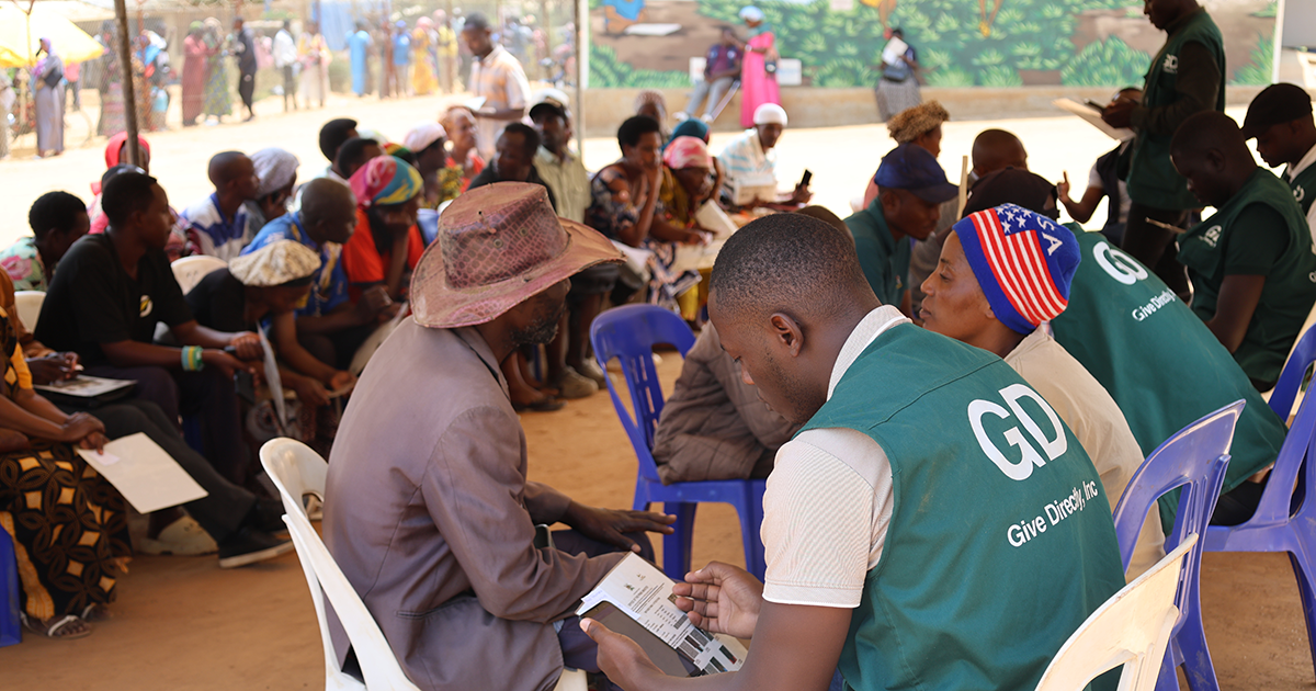 In a shaded public area, a line of people wearing green vests with GiveDirectly logos are each sitting and speaking with someone; a waiting area of other people sit in the background.