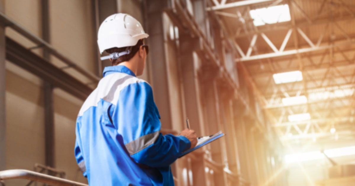 Person in a blue safety uniform and helmet holding a clipboard, looking at an industrial warehouse interior.