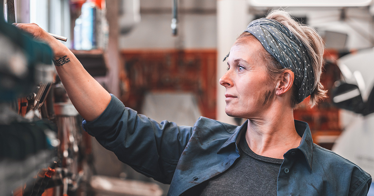 A woman with a patterned headband and a blue work shirt reaches for a tool in a blurred workshop.